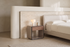 The Bari Bedside Table beside a bed against a travertine panel, with an alabaster table lamp