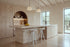 Two Bonassola Counter Stools in white at a travertine kitchen island in a beamed kitchen with olive trees beyond