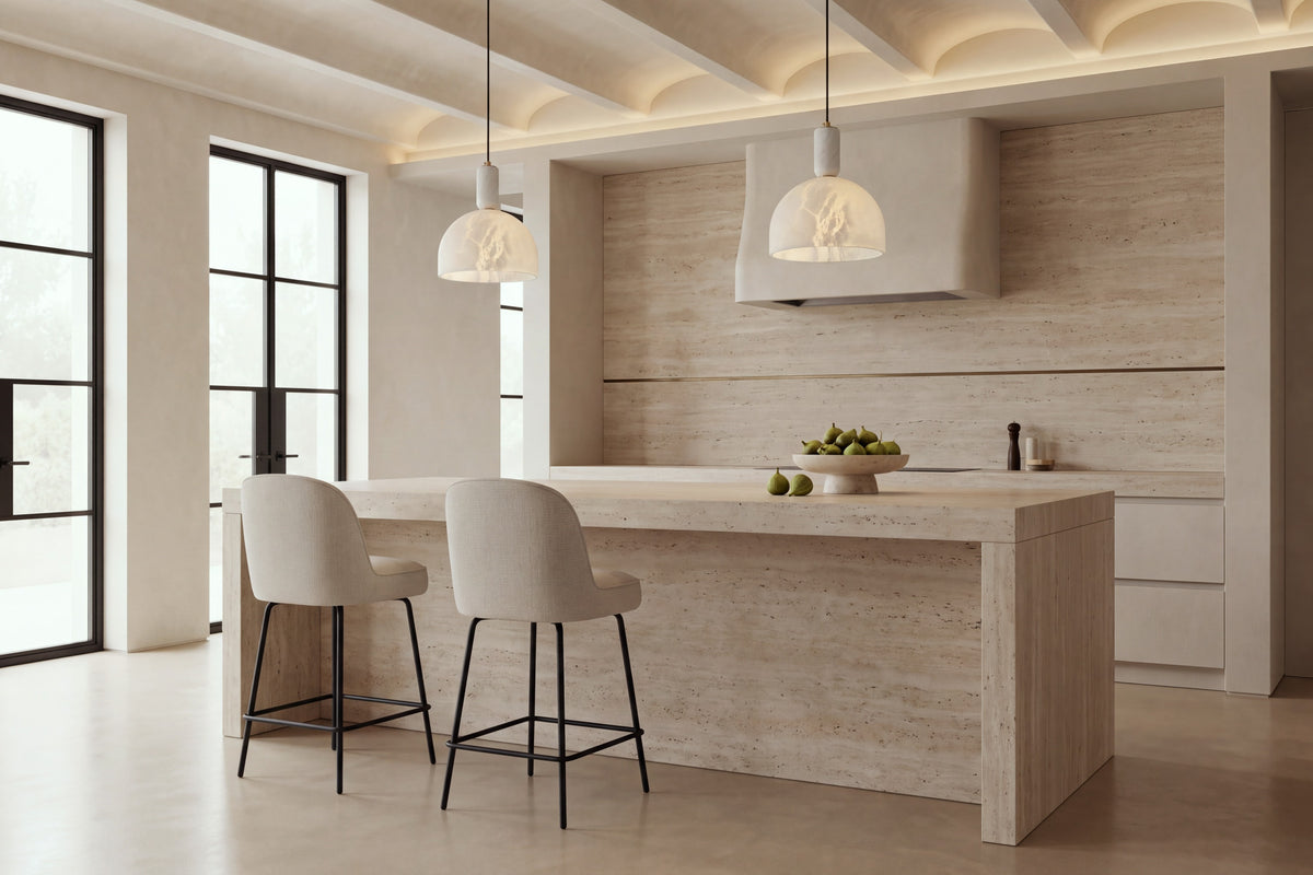 Two Deiva Counter Stools in beige at a travertine kitchen island beneath alabaster pendants