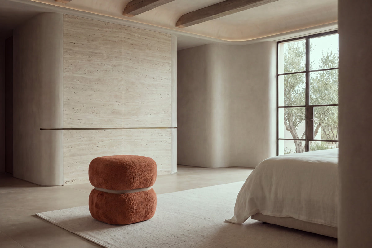 The Gallipoli pouffe in terracotta boucle at the foot of a bed in a calm travertine bedroom