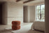 The Gallipoli pouffe in terracotta boucle at the foot of a bed in a calm travertine bedroom