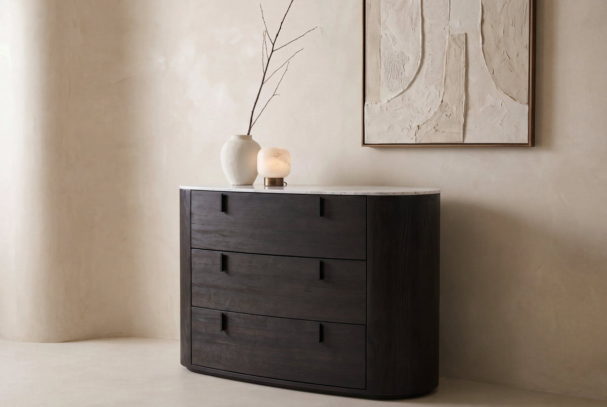 The Bolsena Oval Stone Chest of Drawers against a curved plaster wall with artwork